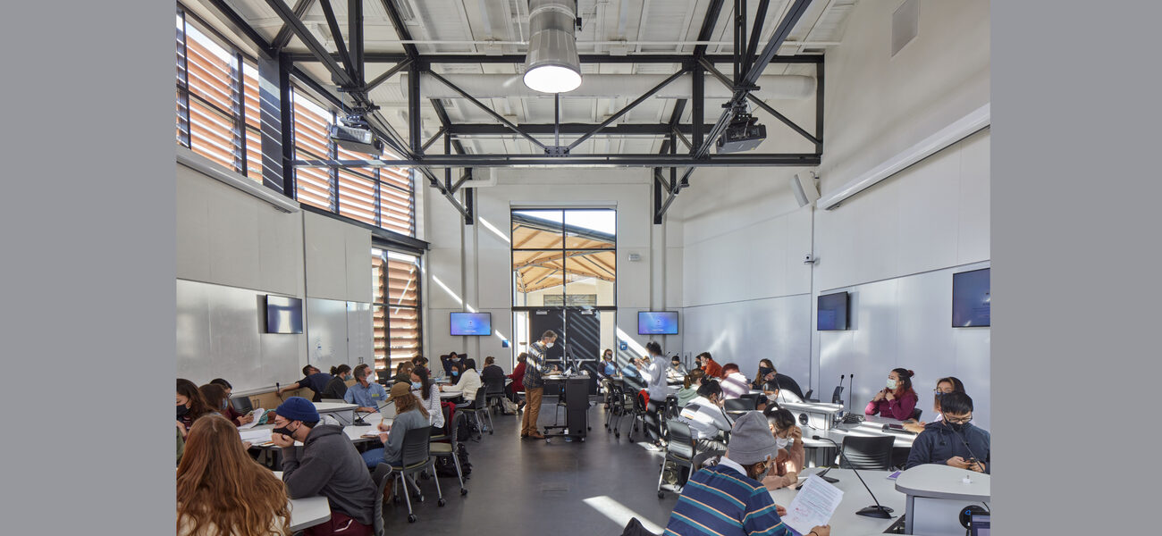 Large classroom with mutiple screens on the walls and students sitting at triangular tables.