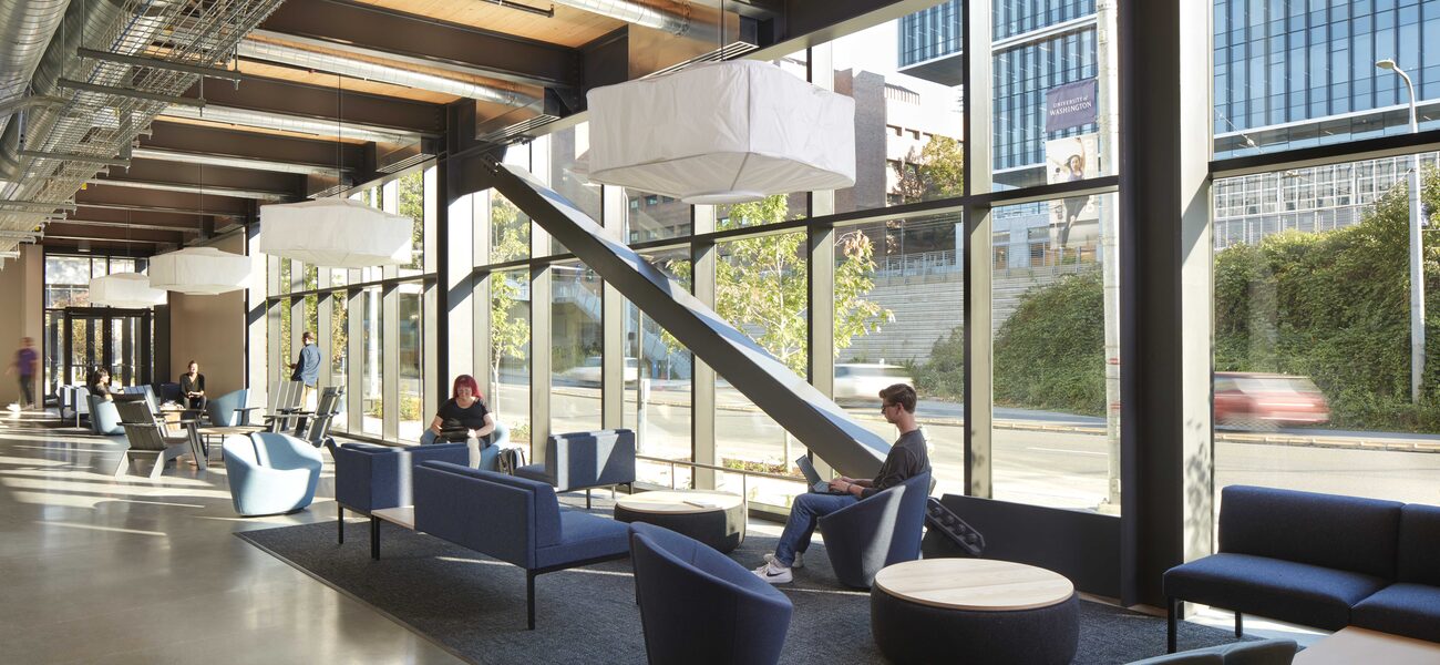 Students sit in comfortable chairs, working on laptops in front of a wall of windows