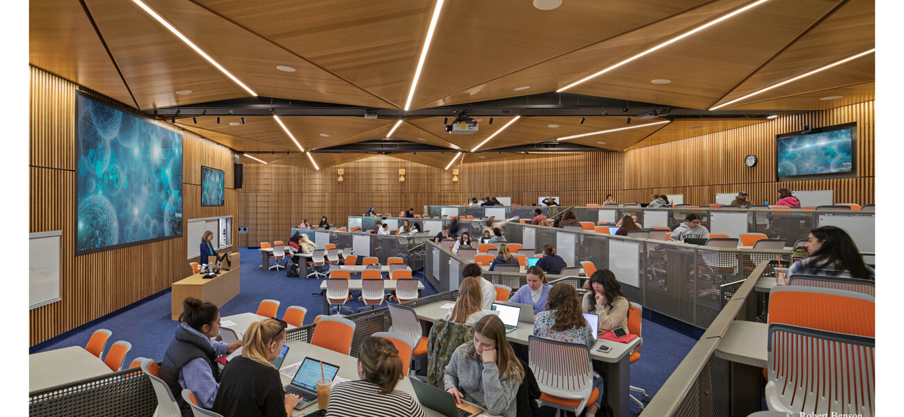 College students sit at six-person tables in a large classroom panelled in wood.