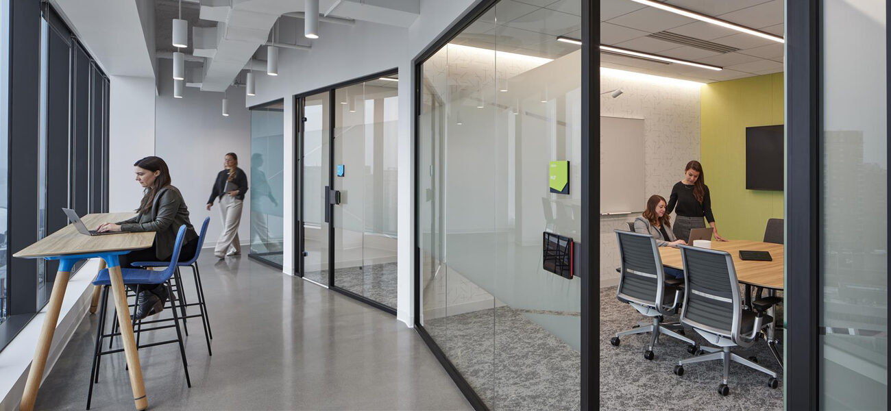 A woman sits at a table in a hallway while two women work in a conference room behind her, separated by a glass wall.