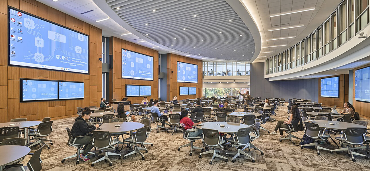 Men and women sit at small circular tables in a large room filled with tables. The walls are lined with large computer screens.