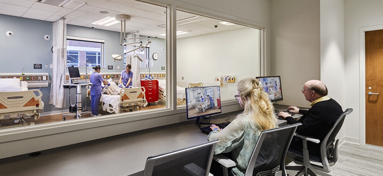 A young man and woman tend to a patient in a hospital bed while an older man and woman observe through a window and on monitors.