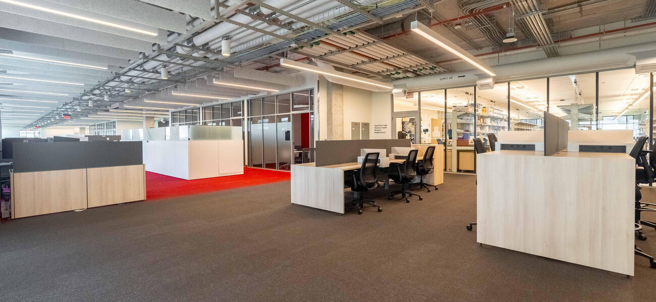 Large room with multiple desks and chairs, and a long glass wall leading to a laboratory. 