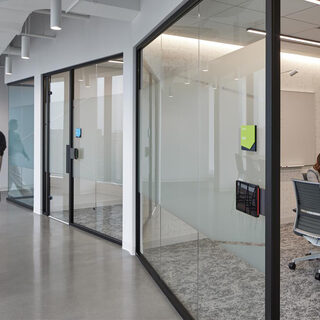 A woman sits at a table in a hallway while two women work in a conference room behind her, separated by a glass wall.